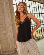 Woman wearing black lace-trim satin cami top with striped trousers, gold bangles, layered necklaces, and oval sunglasses
