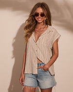 Woman wearing beige striped linen button-up sleeveless shirt with denim cutoff shorts and gold accessories