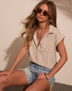 Woman wearing striped linen sleeveless button-down shirt and denim cutoff shorts with gold accessories and tinted sunglasses