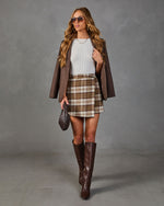 A woman models a brown plaid mini skirt with a white ribbed top, a brown blazer, brown knee-high boots, sunglasses, and a small brown handbag against a gray studio backdrop.