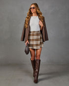 A woman models a brown plaid mini skirt with a white ribbed top, a brown blazer, brown knee-high boots, sunglasses, and a small brown handbag against a gray studio backdrop.