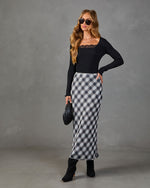 Woman wearing black lace-trim ribbed top, gray plaid satin midi skirt, black heeled boots, holding quilted clutch