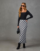 Woman wearing black lace-trim ribbed top, gray plaid satin midi skirt, black heeled boots, holding quilted clutch