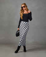 Woman wearing black lace-trim ribbed top, gray plaid midi skirt, black knee-high boots, holding woven bag