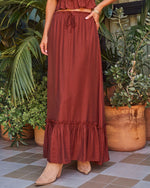 Rust brown maxi skirt with ruffle hem and drawstring waist, paired with tan ankle boots, styled against tropical greenery