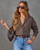 A woman modeling a brown and white vertically striped button-down shirt, styled with blue jeans, a brown belt, a textured brown clutch, and sunglasses against a solid gray studio background.