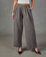 A model wearing a pair of wide-leg, high-waisted gray knit pants with an elastic waistband, paired with a white crop top and dark brown pointed-toe heels, set against a neutral gray studio background.