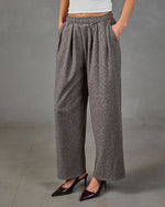 A person wearing loose, wide-leg pleated trousers in a heather gray fabric, paired with a white top and dark brown pointed-toe heels, standing against a gray studio backdrop.