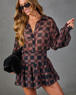 A woman wearing a long-sleeve plaid mini dress in dark red, black, and gray, styled with black sunglasses and holding a black studded hat against a gray studio background.