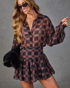 A woman wearing a long-sleeve plaid mini dress in dark red, black, and gray, styled with black sunglasses and holding a black studded hat against a gray studio background.