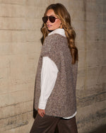 Woman in a brown knit sweater vest over a white button-up shirt, with aviator sunglasses and brown pants against a concrete wall