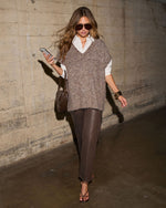 Woman in a brown textured knit sweater vest over a white collared shirt, coated brown pants, and strappy heels, holding a phone
