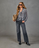 Woman wearing plaid ruffle button-up shirt, gray straight-leg jeans, black ankle boots, holding tan suede clutch