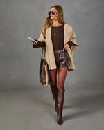 A woman models a stylish fall outfit consisting of a beige open-front coat, a dark brown ribbed knit top, dark brown faux-leather shorts, and dark brown knee-high boots. She is accessorized with a large brown tote bag, sunglasses, and a gold necklace against a gray studio backdrop.