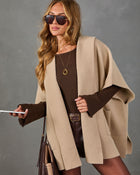 A woman models a beige open-front cape cardigan over a dark brown ribbed long-sleeve top and brown leather pants, accessorized with sunglasses, a gold necklace, and a brown handbag against a textured gray background.