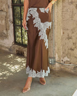 Brown satin maxi skirt with white lace appliqué trim, styled with a gold cuff and clear heels against a rustic concrete wall