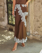 Brown satin maxi skirt with white lace appliqué trim, styled with a gold cuff and clear heels against a rustic concrete wall