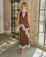 Woman in a brown satin halter top and matching maxi skirt with white lace trim, styled with a woven clutch and clear heels