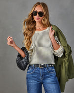Woman wearing cream striped ribbed sweater, olive green jacket, blue jeans, black woven bag, and aviator sunglasses