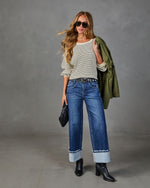 Woman wearing striped knit top, wide-leg cuffed jeans, black ankle boots, holding green utility jacket and quilted clutch