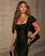 Woman in an olive green velvet midi dress with puff sleeves and corset bustline, styled with gold hoop earrings and bracelets