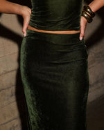 Dark green velvet matching set with fitted crop top and maxi skirt, styled with chunky gold cuff bracelets against a concrete wall