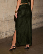 Back view of a dark green velvet maxi skirt with a side slit, styled with black strappy heels and gold cuff bracelets