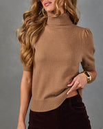 A woman wearing a camel brown short-sleeve mock neck knit sweater with puffed shoulders, paired with dark brown velvet pants, against a solid gray studio background.
