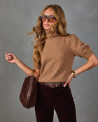 A woman modeling a short-sleeve camel brown knit turtleneck sweater, paired with dark brown corduroy pants, a brown belt, a brown suede shoulder bag, and sunglasses, against a textured gray studio background.