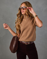 A woman modeling a camel brown knit turtleneck sweater, styled with dark brown velvet pants, a brown leather crossbody bag, and sunglasses, against a textured gray studio background.