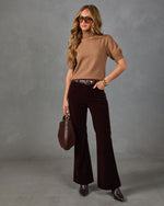 A woman modeling a tan short-sleeve knit top and dark brown wide-leg velvet pants, styled with a brown belt, brown sunglasses, a brown handbag, and brown pointed-toe heels against a textured gray studio background.