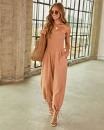 Woman wearing tan strapless smocked jumpsuit with tie-hem ankles, woven tote bag, gold sandals, and sunglasses
