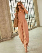 Woman wearing tan smocked strapless jumpsuit with tie ankles, gold heels, woven tote bag, and gold jewelry