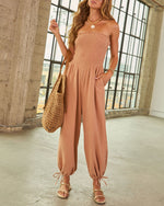 Woman wearing terracotta smocked strapless jumpsuit with tapered legs, woven tote bag, and gold sandals