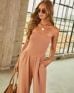 Woman wearing strapless smocked terracotta jumpsuit with wide-leg pants, shell necklace, sunglasses, and woven tote bag