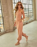 Woman wearing strapless smocked tan jumpsuit with wide legs and tie ankles, styled with gold sandals and hoop earrings