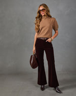 A woman modeling a tan short-sleeve knit turtleneck sweater and dark burgundy velvet flare pants, styled with a brown belt, brown pointed-toe heels, sunglasses, and a brown leather handbag against a textured gray studio backdrop.