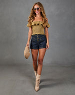 Woman wearing olive smocked off-shoulder crop top, dark denim mini shorts, tan cowboy boots, holding woven clutch