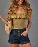 Woman wearing olive green smocked off-shoulder crop top with ruffle trim, dark denim high-waist shorts, and tan woven bag