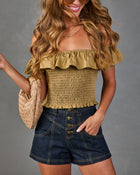 Woman wearing olive green smocked off-shoulder crop top with ruffle trim, dark denim high-waist shorts, and tan woven bag