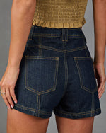 Dark wash high-waist denim shorts with gold contrast stitching and patch pockets, paired with a tan smocked crop top