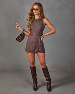 A woman modeling a brown and beige checkered sleeveless romper with matching brown knee-high boots, sunglasses, and a small brown handbag against a gray studio backdrop.