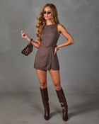 A woman modeling a brown and beige checkered sleeveless romper with matching brown knee-high boots, sunglasses, and a small brown handbag against a gray studio backdrop.