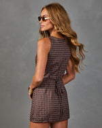 A woman modeling a sleeveless brown and beige gingham mini dress from the back, wearing round sunglasses and gold bracelets against a textured gray studio background.