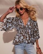 Woman wearing brown floral print boho blouse with bell sleeves, tie front, paired with light wash jeans and tinted sunglasses