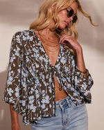 Woman wearing brown floral print tie-front crop blouse with bell sleeves, layered gold necklaces, sunglasses, and light wash jeans
