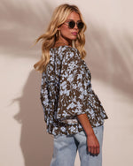 Woman wearing olive floral bell-sleeve blouse with blue flowers and light wash jeans, styled with gold hoop earrings and sunglasses