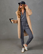 A woman models a cozy two-piece loungewear set consisting of a slate gray zip-up sweatshirt and matching jogger pants, styled with a beige teddy knit cardigan, a navy baseball cap, white sneakers, and sunglasses, while holding a coffee cup against a gray studio background.
