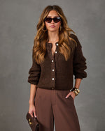Woman wearing a chocolate brown fuzzy knit cardigan with pearl buttons, wide-leg trousers, gold cuff bracelets and cat-eye sunglasses