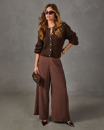 Woman in brown fuzzy button-up cardigan, wide-leg chocolate trousers, cat-eye sunglasses and pointed heels holding a clutch